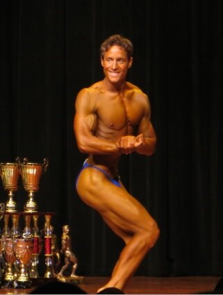 Smiling bodybuilder in blue trunks doing a side chest pose beside gold trophies on stage