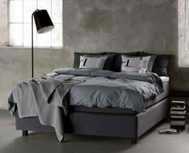 Modern grey bed with charcoal bedding against a concrete wall, black lamp and stacked books nearby