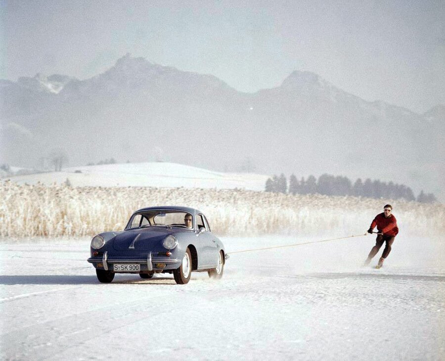 Vintage light-blue Porsche on snow towing a skier in a red jacket toward hazy mountains