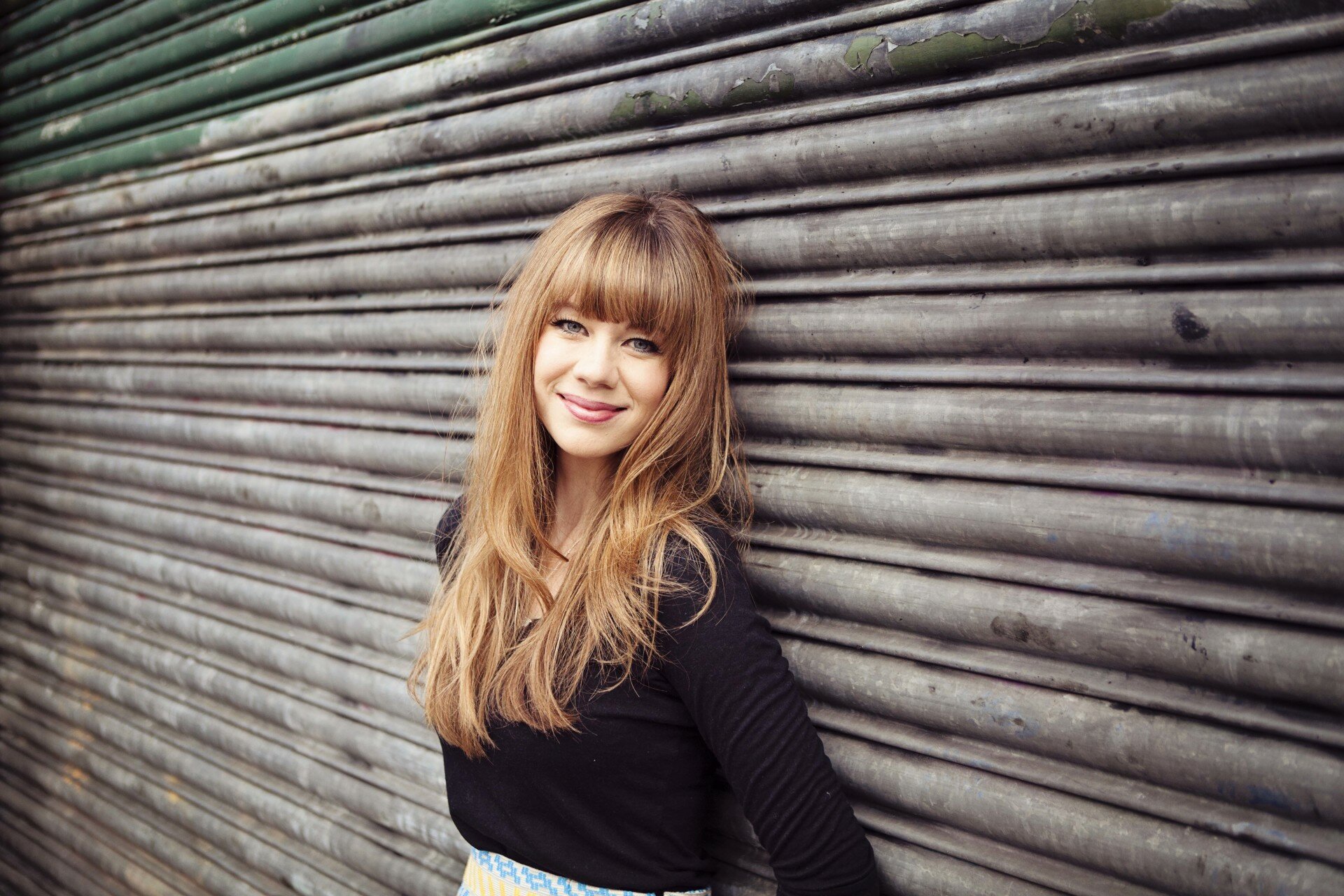 Young woman with strawberry-blonde bangs smiling while leaning against a grey corrugated metal door