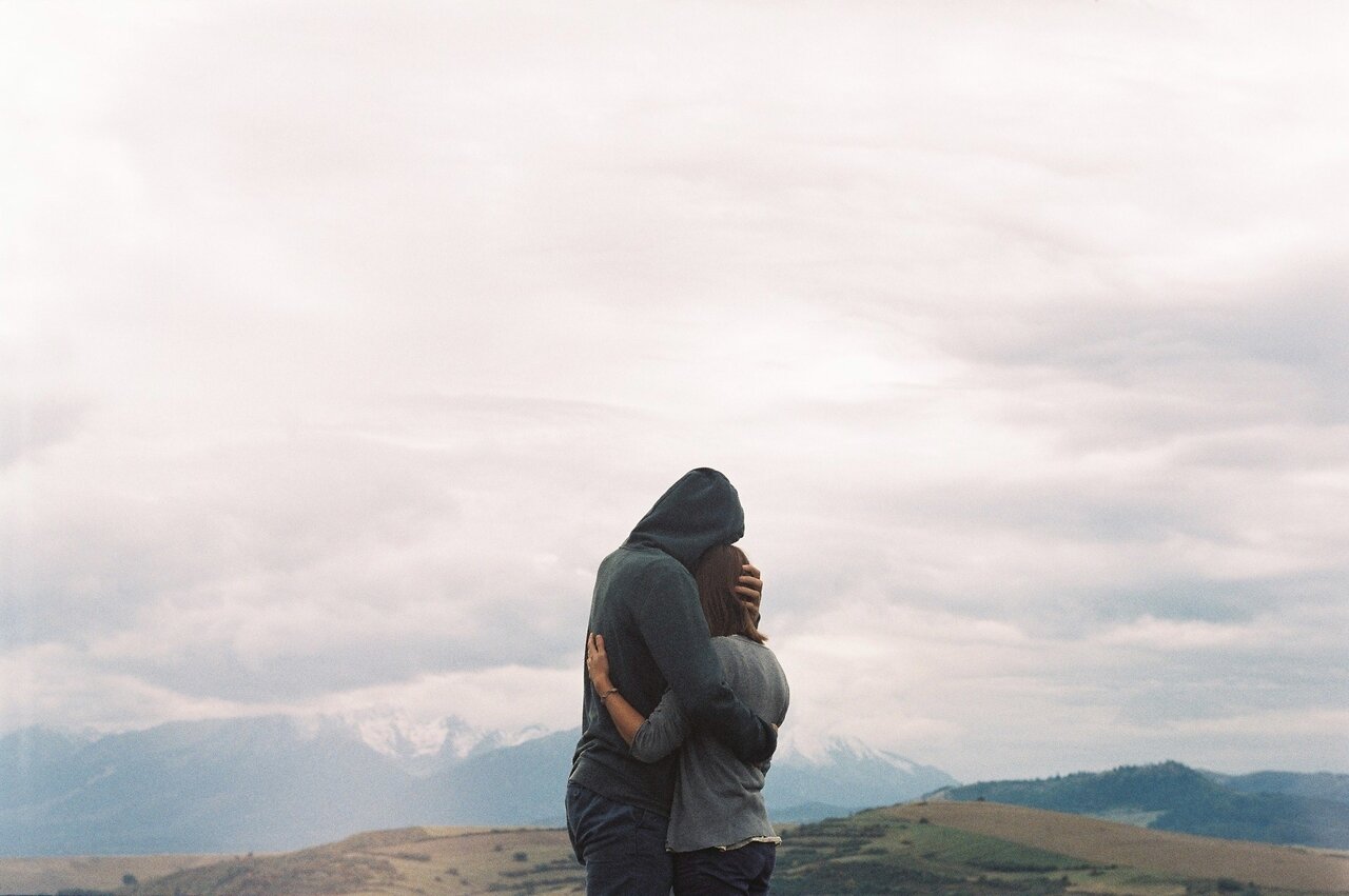 Couple embracing on a hillside, man in a dark hoodie holding a woman against distant snowy peaks