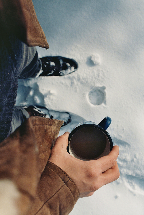 First-person view holding a blue enamel coffee mug in deep snow, brown jacket and snowy black boots