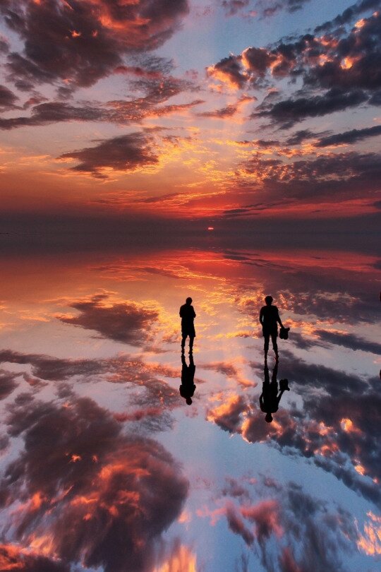 Two silhouettes on a flooded salt flat reflecting an orange and purple sunset sky