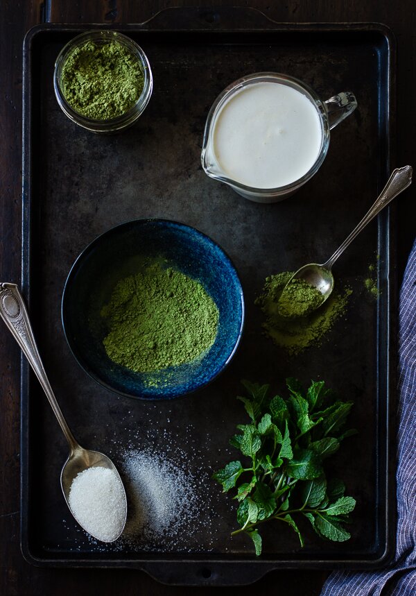 Flat lay of matcha powder, milk pitcher, sugar spoon, and fresh mint on a dark rustic tray