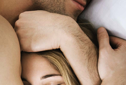 Overhead close-up of a stubbled man embracing a blonde woman resting on a white pillow