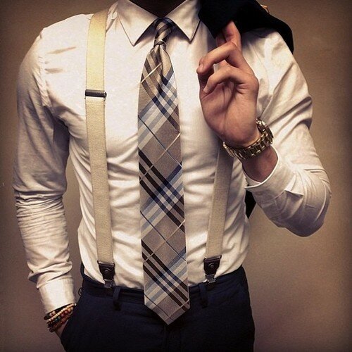 Man in a white shirt, plaid tie, and tan suspenders holding a dark jacket over his shoulder